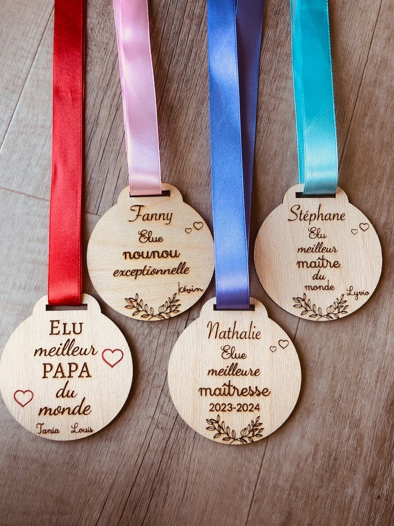May include: Four wooden medallions with ribbons, each with a different inscription. The first medallion says "Elu meilleur Papa du monde" with hearts and the names "Tania Louis". The second medallion says "Fanny Elue nounou exceptionnelle" with hearts and the name "K&eacute;vin". The third medallion says "Nathalie Elue meilleure ma&icirc;tresse 2023-2024" with hearts. The fourth medallion says "St&eacute;phane Elu meilleur ma&icirc;tre du monde" with hearts and the name "Lyvio".