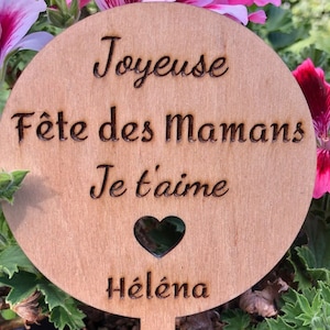 Peut inclure: Une pancarte en bois avec le texte "Joyeuse Fête des Mamans Je t'aime" et une découpe en forme de cœur. Le texte "Hélène" est en bas de la pancarte.