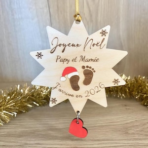 Puede incluir: Adorno navideño de madera en forma de estrella con el texto "Joyeux Noël Papy et Mamie" y "J'arrive en 2026". Presenta un gorro de Papá Noel, huellas y un dije de corazón rojo. Incluye una cuerda dorada para colgar.