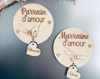 May include: Two round wooden plaques with the phrases "Parrain d'amour" and "Marraine d'amour" engraved in a decorative font. Each plaque features a heart-shaped charm with a name. The plaques are light brown with a heart and hand-holding design.