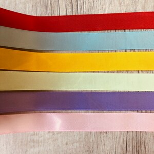 May include: Six different colored satin ribbons arranged horizontally on a woodgrain surface. The ribbons are red, light blue, yellow, light green, purple, and pink.