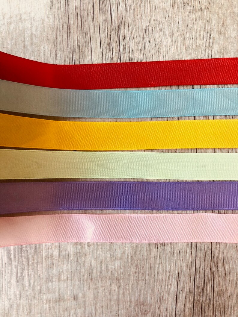 May include: Six different colored satin ribbons arranged horizontally on a woodgrain surface. The ribbons are red, light blue, yellow, light green, purple, and pink.