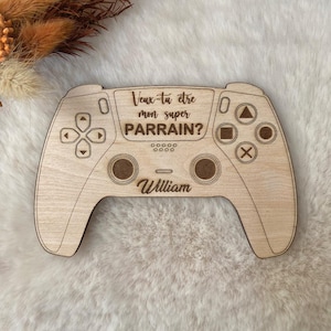 Può includere: Un controller di gioco in legno con testo inciso. Il testo recita "Veux-tu être mon super PARRAIN?" e "William". Il controller ha pulsanti e pad direzionali. Il legno è di colore marrone chiaro.