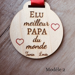 May include: A wooden medal with the text "Elu meilleur PAPA du monde" and two red hearts. The names "Tania" and "Louis" are written below the text. The medal is attached to a red ribbon. The text "Mod&egrave;le 2" is written below the medal.