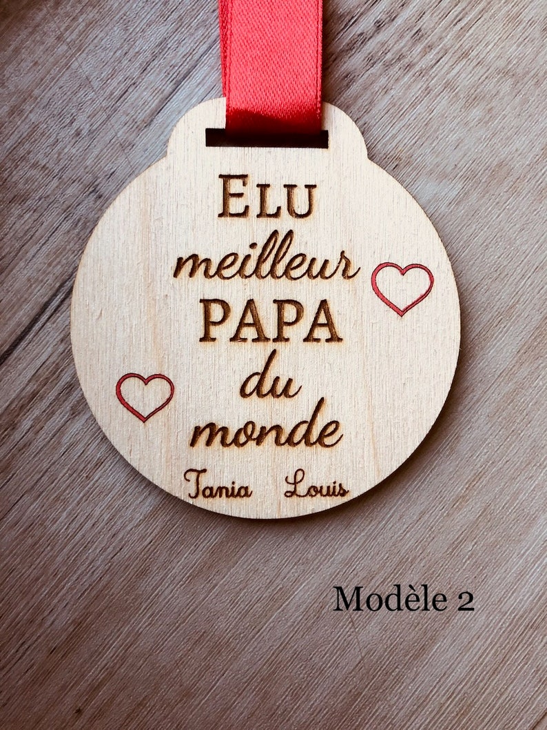 May include: A wooden medal with the text "Elu meilleur PAPA du monde" and two red hearts. The names "Tania" and "Louis" are written below the text. The medal is attached to a red ribbon. The text "Mod&egrave;le 2" is written below the medal.