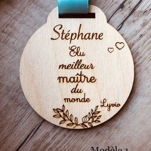 May include: A wooden round medal with a blue ribbon. The medal has the text "St&eacute;phane Elu meilleur ma&icirc;tre du monde" and two small hearts. The text "Lyrio" is at the bottom of the medal. The text "Mod&egrave;le 1" is written on the wooden surface below the medal.