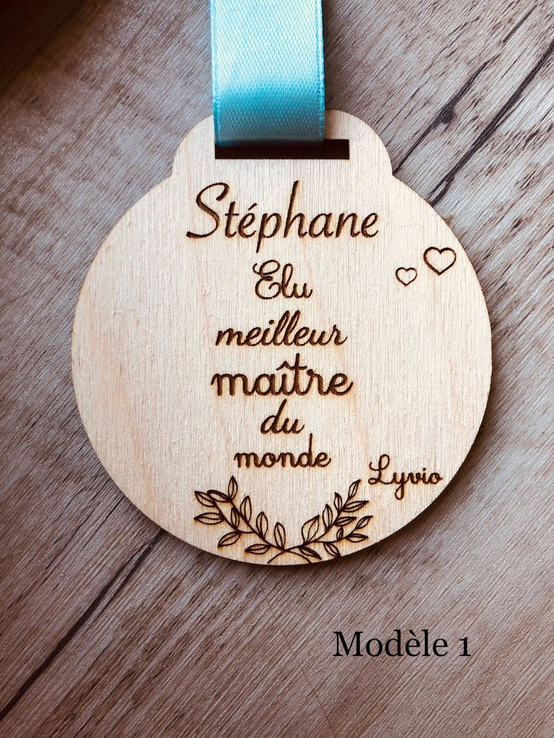 May include: A wooden round medal with a blue ribbon. The medal has the text "St&eacute;phane Elu meilleur ma&icirc;tre du monde" and two small hearts. The text "Lyrio" is at the bottom of the medal. The text "Mod&egrave;le 1" is written on the wooden surface below the medal.