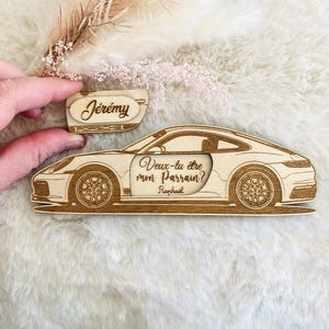 Peut inclure: Objet décoratif en bois en forme de Porsche, gravé avec le nom "Jérémy" et le texte "Veux-tu être mon Parrain? Raphael". La voiture, de couleur marron clair, présente des détails précis et un design élégant, parfait pour un baptême.