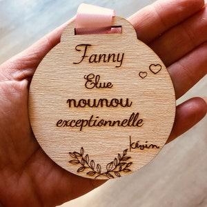 May include: A round wooden plaque with the text "Fanny Elue nounou exceptionnelle Kevin" and two small hearts. The plaque has a pink ribbon attached to the top.