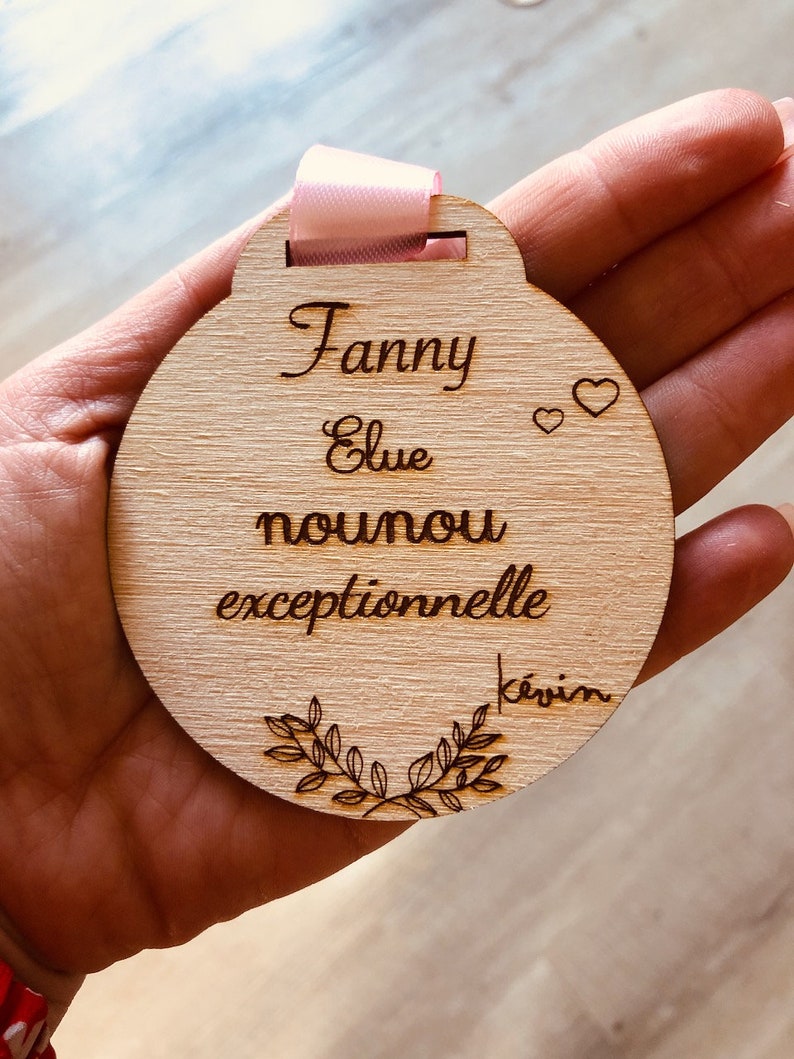 May include: A round wooden plaque with the text "Fanny Elue nounou exceptionnelle Kevin" and two small hearts. The plaque has a pink ribbon attached to the top.