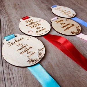 May include: Four wooden medallions with ribbons, each with a different inscription. The medallions are engraved with the words "Elu meilleur Papa du monde", "Elu meilleur ma&icirc;tre du monde", "Nathalie", and "Fanny, la nounou exceptionnelle".