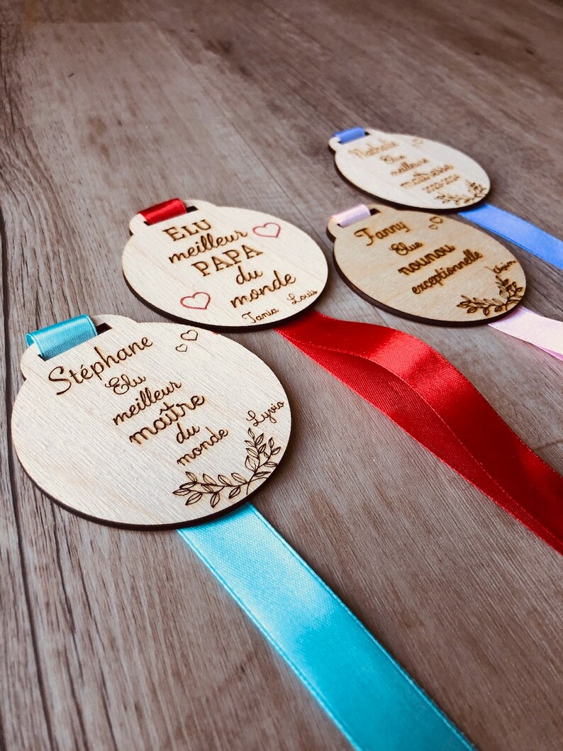 May include: Four wooden medallions with ribbons, each with a different inscription. The medallions are engraved with the words "Elu meilleur Papa du monde", "Elu meilleur ma&icirc;tre du monde", "Nathalie", and "Fanny, la nounou exceptionnelle".