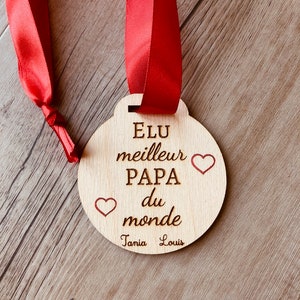 May include: A wooden medal with a red ribbon, engraved with the text "Elu meilleur Papa du monde" and two hearts. The names "Tania" and "Louis" are also engraved on the medal.
