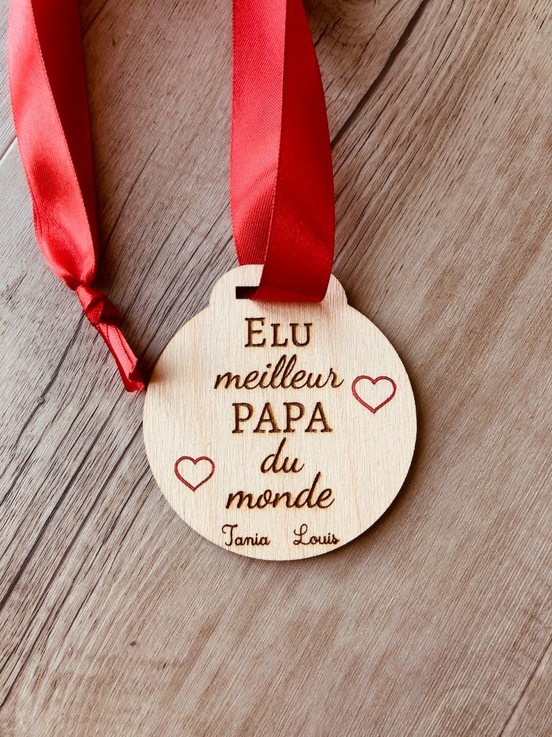 May include: A wooden medal with a red ribbon, engraved with the text "Elu meilleur Papa du monde" and two hearts. The names "Tania" and "Louis" are also engraved on the medal.