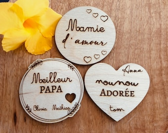 May include: Three wooden plaques with engraved messages. The first plaque is round and says "Maman d'amour" with hearts. The second plaque is round and says "Meilleur Papa" with a heart and the names "Olivia" and "Mathias". The third plaque is heart-shaped and says "Nounou adorée" with the name "Anna" and the word "Tom".