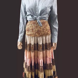 May include: A long, multi-coloured skirt with a tiered design. The skirt is made of different fabrics and has a brown, beige, pink, and red colour scheme. The skirt is worn with a light blue button-down shirt with white polka dots.