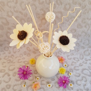 Op de afbeelding: Een witte keramische vaas met een ronde vorm bevat een boeket van houten stokjes met decoratieve ballen en bloemen. De vaas is omringd door kleine witte, roze en gele bloemen op een beige, getextureerd oppervlak.