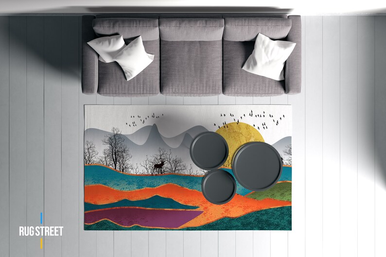 May include: A gray sectional sofa with white pillows sits on a white wood floor. A colorful rug with a mountain and sun design is placed in front of the sofa. Three black round coffee tables are on the rug.
