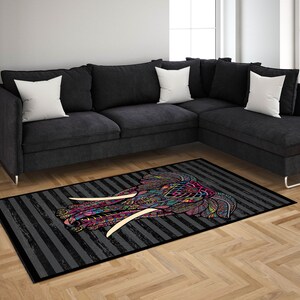 May include: A colorful patterned elephant head rug with black and white stripes on a gray background. The rug is placed in front of a black sectional sofa with white pillows.