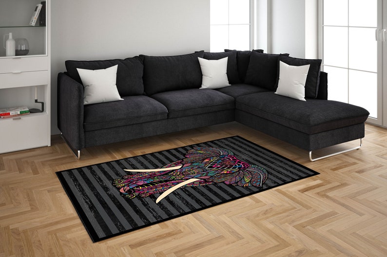 May include: A colorful patterned elephant head rug with black and white stripes on a gray background. The rug is placed in front of a black sectional sofa with white pillows.