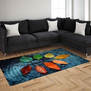 May include: A blue and brown rug with a design of colorful autumn leaves on a wooden background. The rug is placed in front of a gray sectional sofa with white pillows.