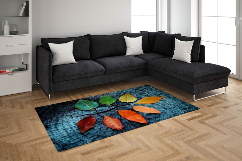 May include: A blue and brown rug with a design of colorful autumn leaves on a wooden background. The rug is placed in front of a gray sectional sofa with white pillows.