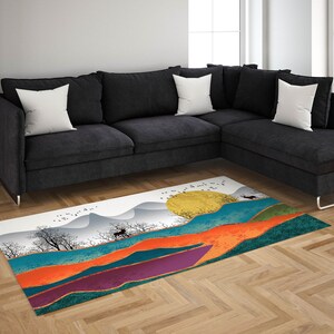 May include: A colorful abstract rug with a white background featuring a stylized landscape with mountains, a sun, and a deer. The rug has shades of orange, purple, teal, and gold.