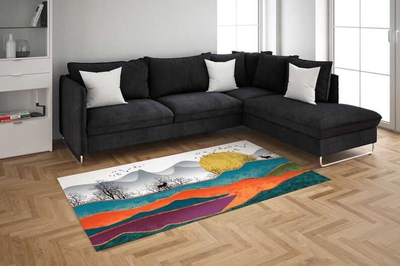 May include: A colorful abstract rug with a white background featuring a stylized landscape with mountains, a sun, and a deer. The rug has shades of orange, purple, teal, and gold.