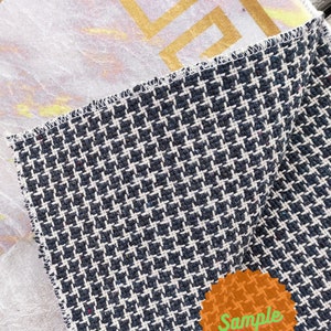 May include: A close-up of a black and white patterned fabric with a gold and white patterned fabric underneath. The fabric has a "Sample" label on it.