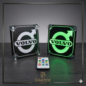 Puede incluir: Se muestran dos luces LED cuadradas negras con el logotipo de Volvo, una blanca y otra verde. Debajo hay un mando a distancia con botones de colores. Las luces están fijadas con ventosas. El logotipo de Barjoe Print está en la parte inferior.