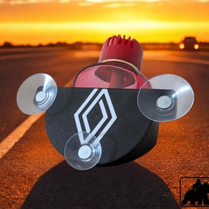 Könnte beinhalten: Ein Auto-Lufterfrischer mit schwarzer Basis und weißem Rautenmuster. Der Lufterfrischer hat einen roten Deckel und wird mit drei Saugnäpfen an der Windschutzscheibe befestigt. Der Hintergrund ist eine Straße bei Sonnenuntergang.