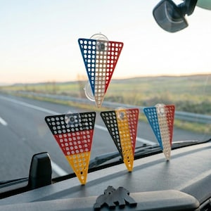 May include: Four triangular car flags with suction cups, featuring the colors of different national flags. The flags are red, white, and blue; black, red, and yellow; and black, yellow, and red. The flags are on a car dashboard.