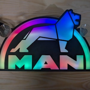 May include: A black and white MAN logo with a lion silhouette. The logo is illuminated with a rainbow of colours. The logo is mounted on a black background with two suction cups.