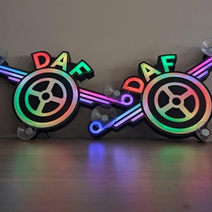May include: Two illuminated DAF logo signs with rainbow-colored lights. Each sign features a circular design with the letters DAF in red, green, and blue. The signs have suction cups for mounting and are set against a neutral background.