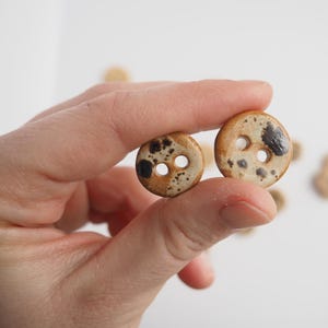 Può includere: Due piccoli bottoni in ceramica rotondi a due fori, con una base color marrone chiaro e motivi a macchie marrone scuro. I bottoni hanno una finitura liscia e lucida, sono di forma circolare e presentano due fori centrali per il fissaggio.
