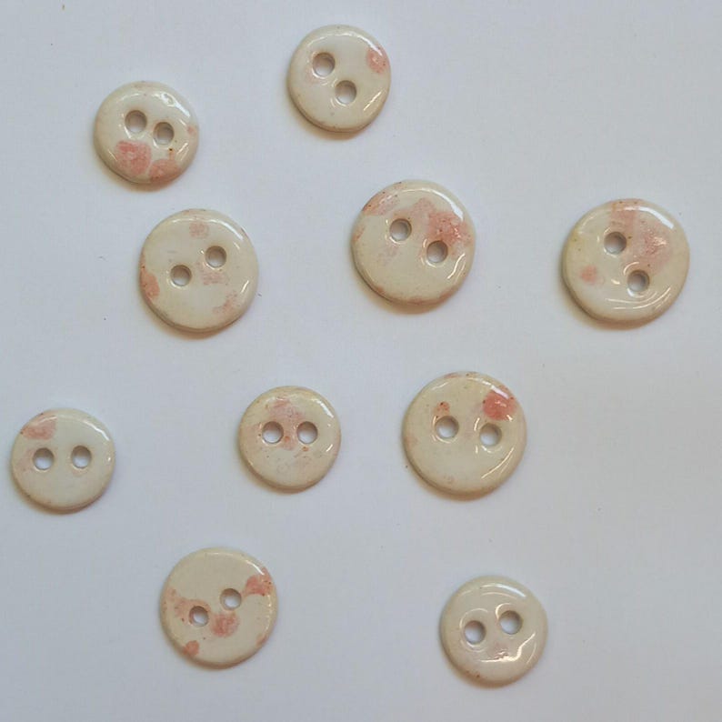 May include: Ten white ceramic buttons with two holes each. The buttons have a speckled pink and white glaze.