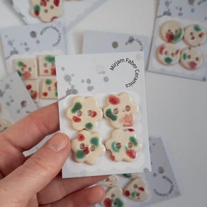 Peut inclure: Gros plan sur des boutons en céramique de différentes formes et tailles, chacun avec un design unique. Les boutons sont blanc cassé avec des accents rouges et verts. Le texte "Mirjam Faber Ceramics" est visible sur l'emballage.