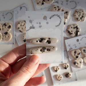 Puede incluir: Botones y cierres de cerámica crema con motas negras, de varias formas. Los botones están empaquetados en tarjetas blancas con el texto "Mirjam Faber Ceramics".