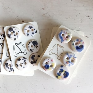 Può includere: Set di bottoni decorativi su cartoncini bianchi. Alcuni bottoni hanno un motivo maculato bianco e nero, mentre altri presentano disegni astratti colorati in blu, rosa e giallo. Ogni cartoncino ha anche una piccola illustrazione di un maglione.