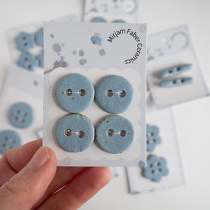 May include: A selection of ceramic buttons in different shapes and sizes. Four round, pale blue buttons with two holes are displayed on a white card, labelled "Mirjam Faber Ceramics". Other cards with buttons are visible in the background, with a hand holding the card.