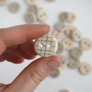 May include: A close-up of a white ceramic button with a black grid pattern and two holes. The button is being held by a hand.