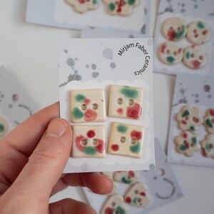 May include: A hand holding a card with four square ceramic buttons. The buttons are off-white with red and green accents. Other cards with ceramic buttons in various shapes and colours are in the background. The card reads "Mirjam Faber Ceramics."