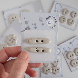 May include: A hand holds a card of two off-white ceramic toggle buttons with brown speckles. Other cards show various ceramic buttons, including round and flower-shaped designs. The cards have the text "Mirjam Faber Ceramics".