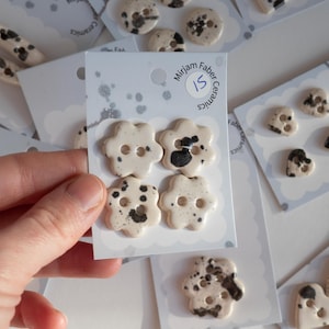May include: Four off-white ceramic buttons with black speckled patterns, shaped like six-petaled flowers, each featuring two central holes. The buttons are mounted on a white display card labeled Mirjam Faber Ceramics with the number 15.