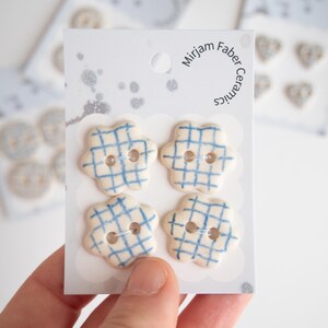 May include: Four off-white ceramic buttons with a blue grid pattern, shaped like six-petaled flowers. Each button has two central holes. The buttons are attached to a white display card labelled Mirjam Faber Ceramics with a circular hole at the top.