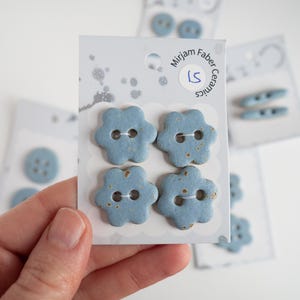 May include: Four light blue ceramic flower-shaped buttons with brown speckles are displayed on a white card. The card has the text "Mirjam Faber Ceramics" and a sticker with "LS". Other cards with similar buttons are in the background.
