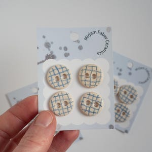 Può includere: Quattro bottoni in ceramica color crema con un motivo a griglia blu, presentati su una carta azzurra. La carta ha un bordo smerlato e il testo "Mirjam Faber Ceramics".
