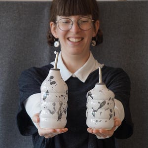 May include: Two white ceramic oil and vinegar bottles with black splatter paint designs. The bottles have silver tops and are held by a person wearing a black shirt and white shirt underneath.