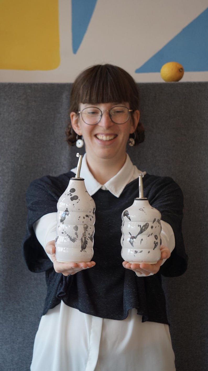 May include: Two white ceramic oil and vinegar bottles with black splatter paint designs. The bottles have silver tops and are held by a person wearing a black shirt and white shirt underneath.