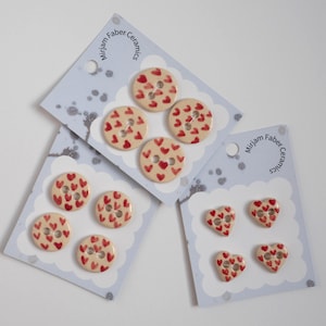 Peut inclure: Trois cartes de présentation marquées Mirjam Faber Ceramics, contenant des boutons en céramique avec des motifs de cœurs rouges. Deux cartes présentent quatre boutons ronds, tandis que la troisième affiche quatre boutons en forme de cœur.
